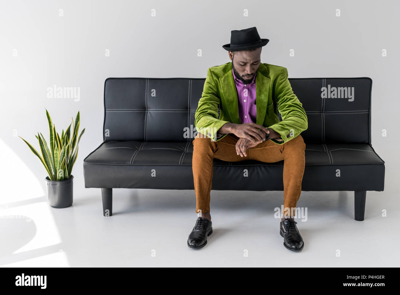 Moda hombre afroamericano en hat control de tiempo mientras está sentado en un sofá negro Foto de stock