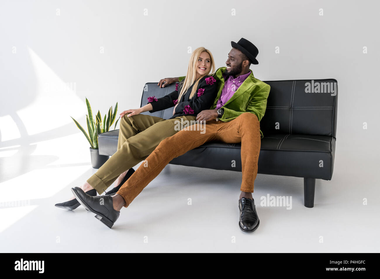 Pareja de moda multicultural alegre descansando sobre sofás negros sobre fondo gris Foto de stock