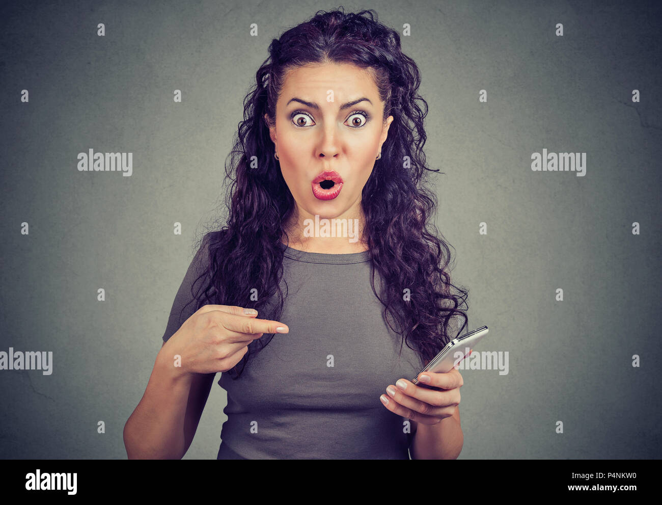 Mujer joven mirando con expresión de terror en la cámara apuntando al teléfono inteligente, después de leer noticias alarmantes sobre la página web aislado sobre fondo gris. Foto de stock