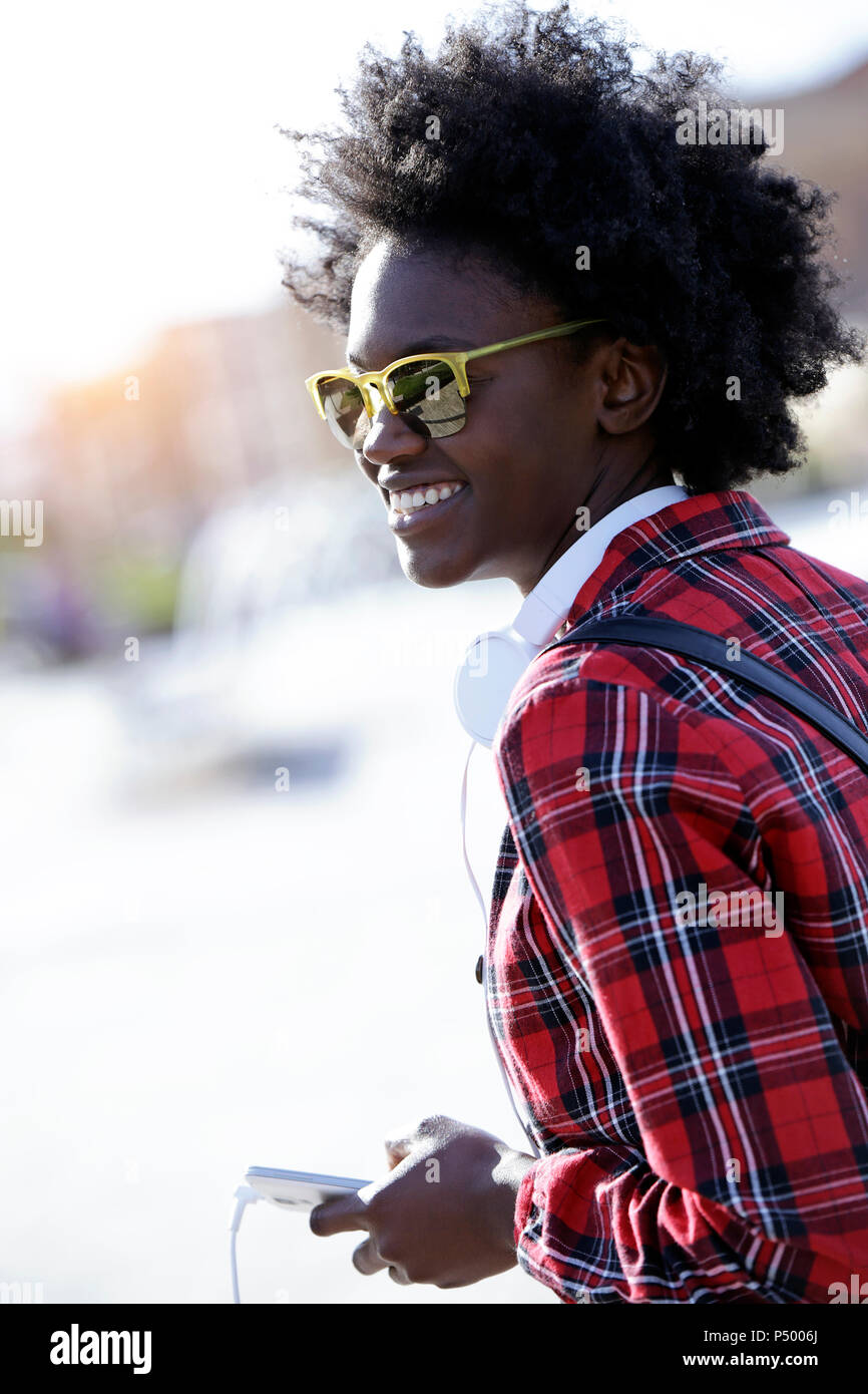 Retrato de mujer sonriente con auriculares y smartphone usando gafas de sol reflejados Foto de stock