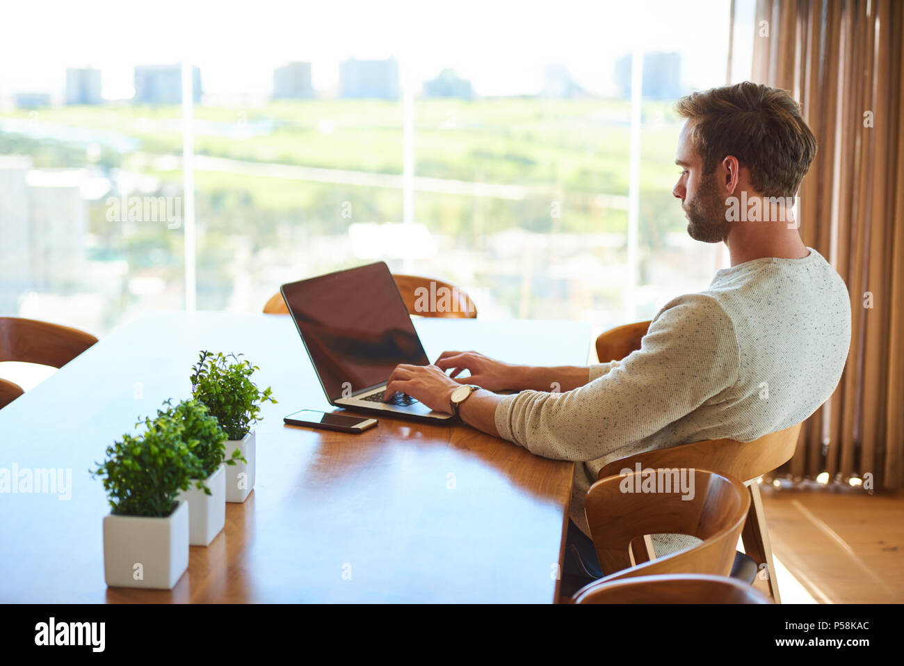 Día moderno hombre blanco ocupado trabajando en su portátil mientras está  sentado en su mesa de comedor en casa, con una preciosa vista de la ciudad  a través de las grandes ventanas