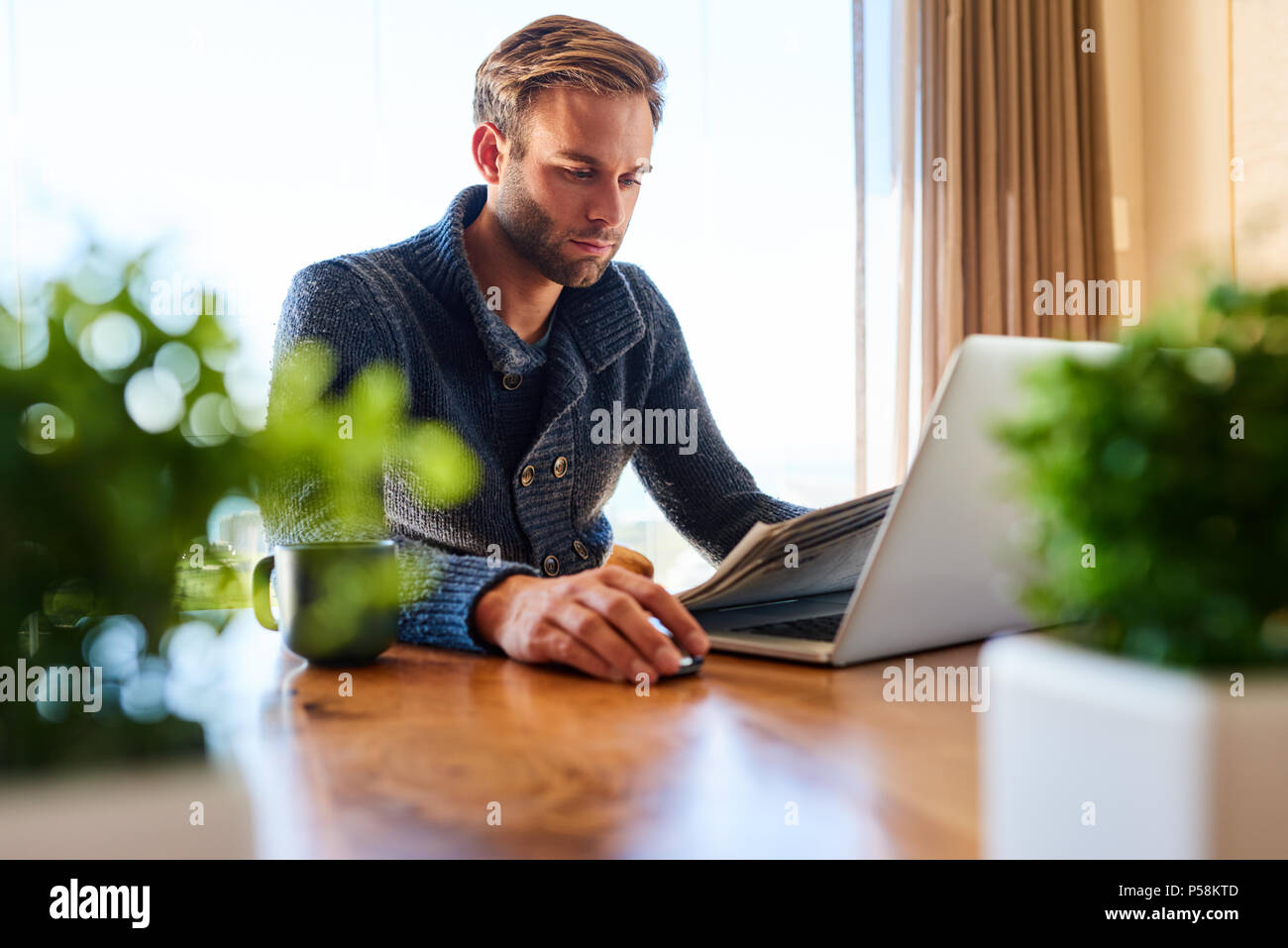 Apuesto hombre moderno ocupado leyendo el periódico y comprobando sus mails para ponerse al día de la actualidad desde la comodidad de su hogar mientras está sentado en su mesa de comedor. Foto de stock