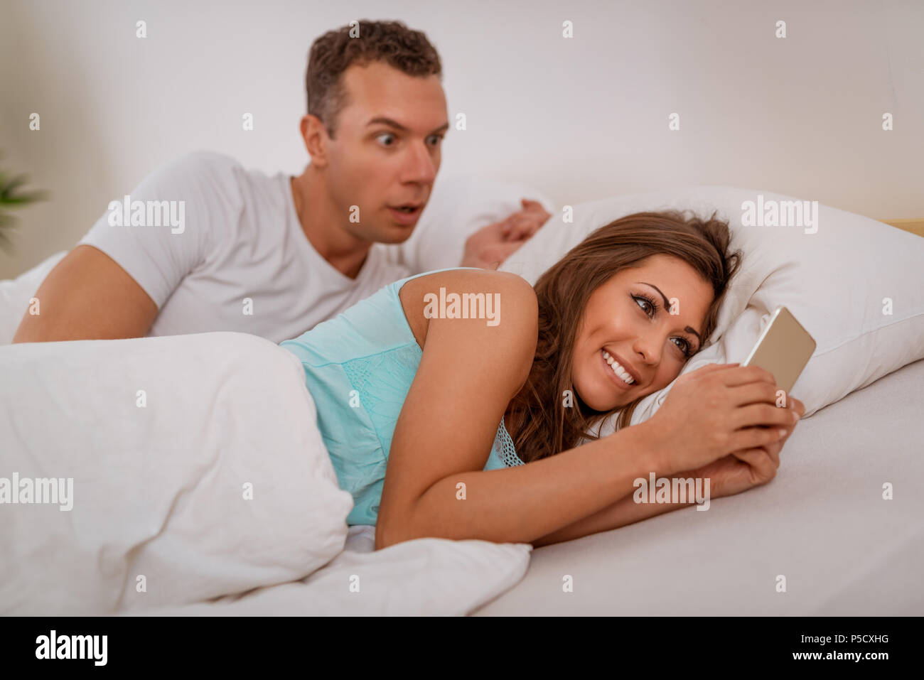 Esposa engañando a su marido a través de teléfono móvil en la cama, el marido se engancha a ella Fotografía de stock