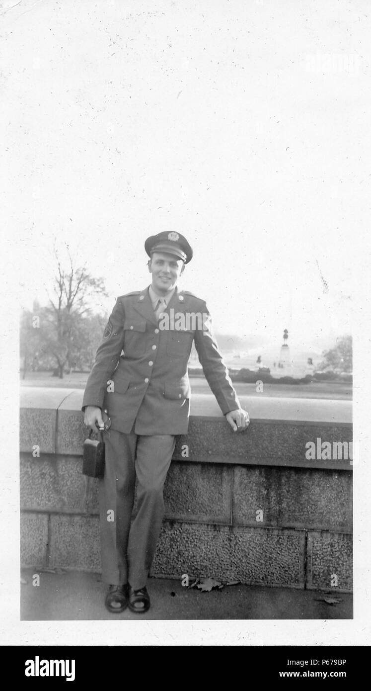 Fotografía en blanco y negro, mostrando un soldado masculino, de longitud completa, que miran a la cámara y sonriendo, vistiendo un uniforme y gorra, sosteniendo un estuche de cámara, y se apoya en un muro de piedra bajo con un parque y monumentos en el fondo, probablemente fotografiado en Ohio durante la II Guerra Mundial, en 1945. () Foto de stock