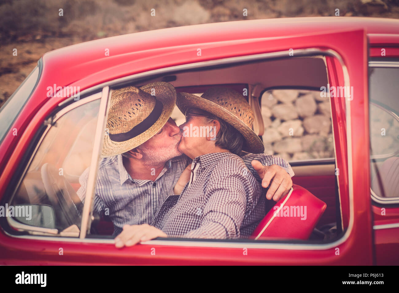 Pareja de ancianos con sombrero, con gafas, con el pelo blanco y gris, con camisa casual, en vintage coche rojo en vacaciones disfrutando de tiempo y vida. Con un chee Foto de stock