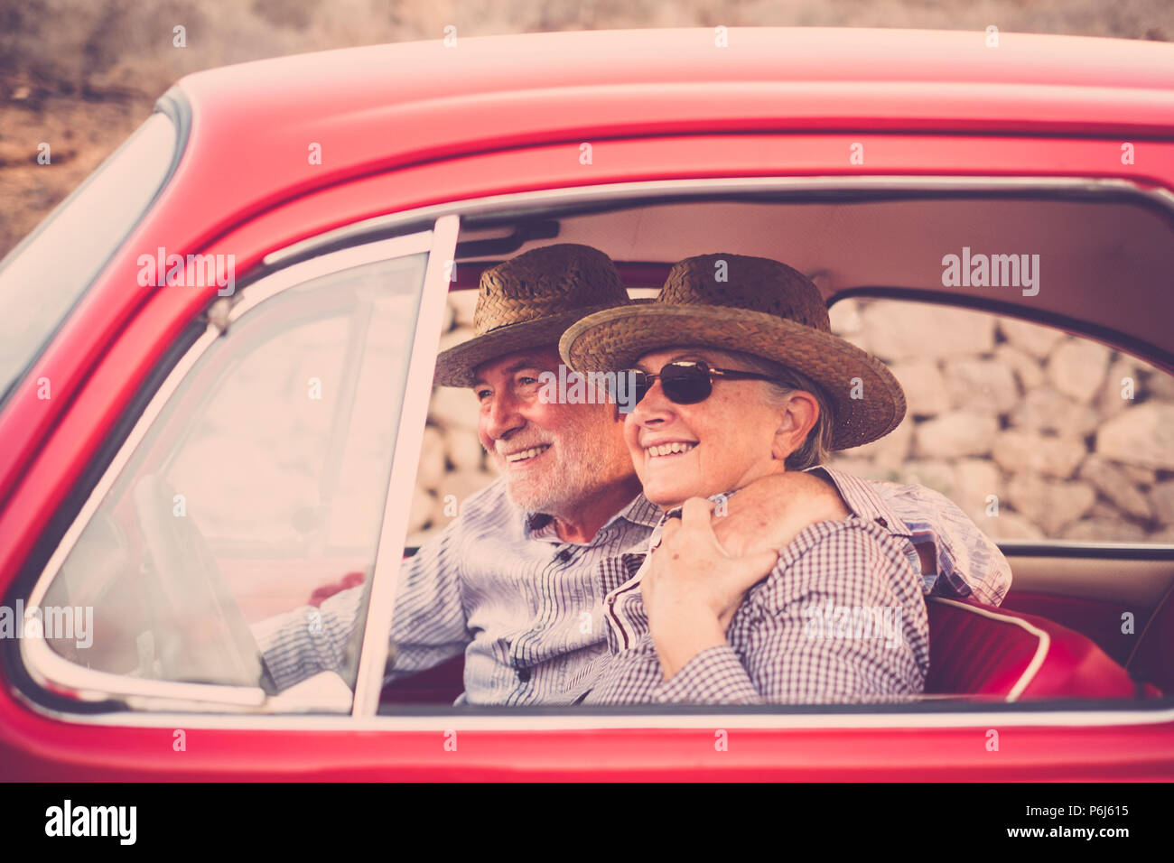 Pareja de ancianos con sombrero, con gafas, con el pelo blanco y gris, con camisa casual, en vintage coche rojo en vacaciones disfrutando de tiempo y vida. Con un chee Foto de stock