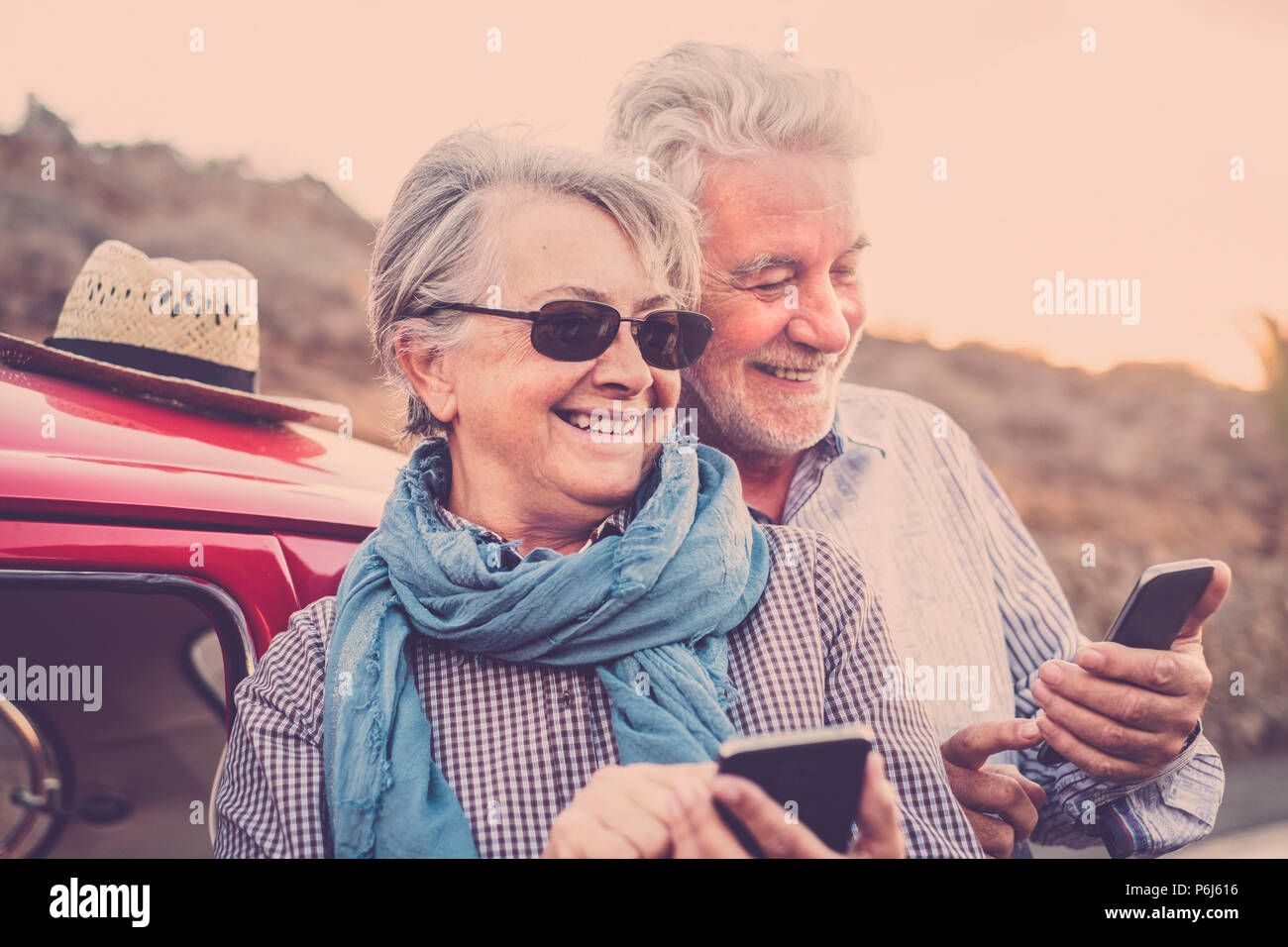 Pareja de ancianos con sombrero, con gafas, con el pelo blanco y gris, con camisa casual, en vintage coche rojo en vacaciones disfrutando de tiempo y vida. Con un chee Foto de stock