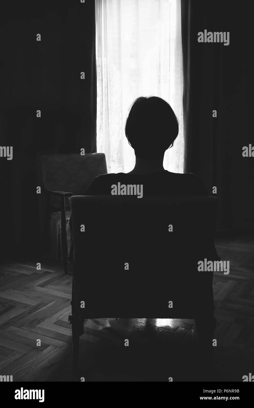 Mujer sentada sola en una sala oscura mirando a través de una ventana. Blanco y negro. Foto de stock