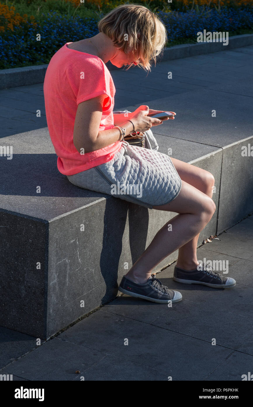 Mujer joven con cabello rubio sentado en un banco de un parque en la tarde el Sol trabajando en su teléfono móvil en la Place de l'Hotel de Ville St Quentin Francia Foto de stock