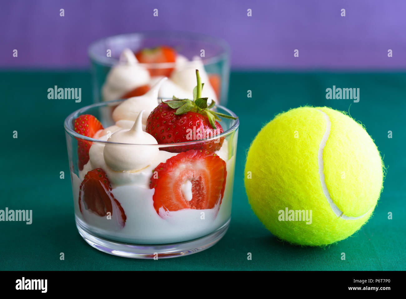Wimbledon inspirado de crema batida, Merengues y fresas frescas en un recipiente de cristal sobre un fondo de color verde y violeta con una pelota de tenis Foto de stock