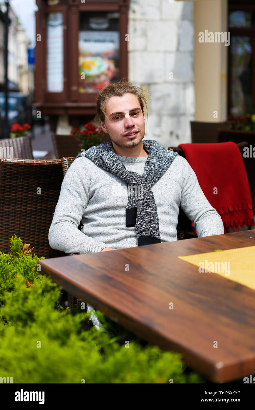 Hombre joven sentado en la cafetería de la calle cerca de las plantas verdes, vistiendo un suéter gris. Foto de stock