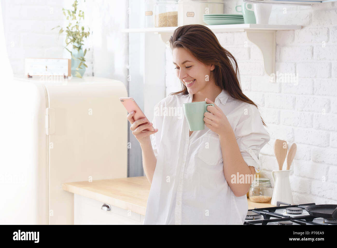 Mujer joven con inclinada del smartphone en la mesa de la cocina con la taza de café y el organizador en un hogar moderno. Mujer sonriente leer mensaje en el teléfono. Morena ha. Foto de stock