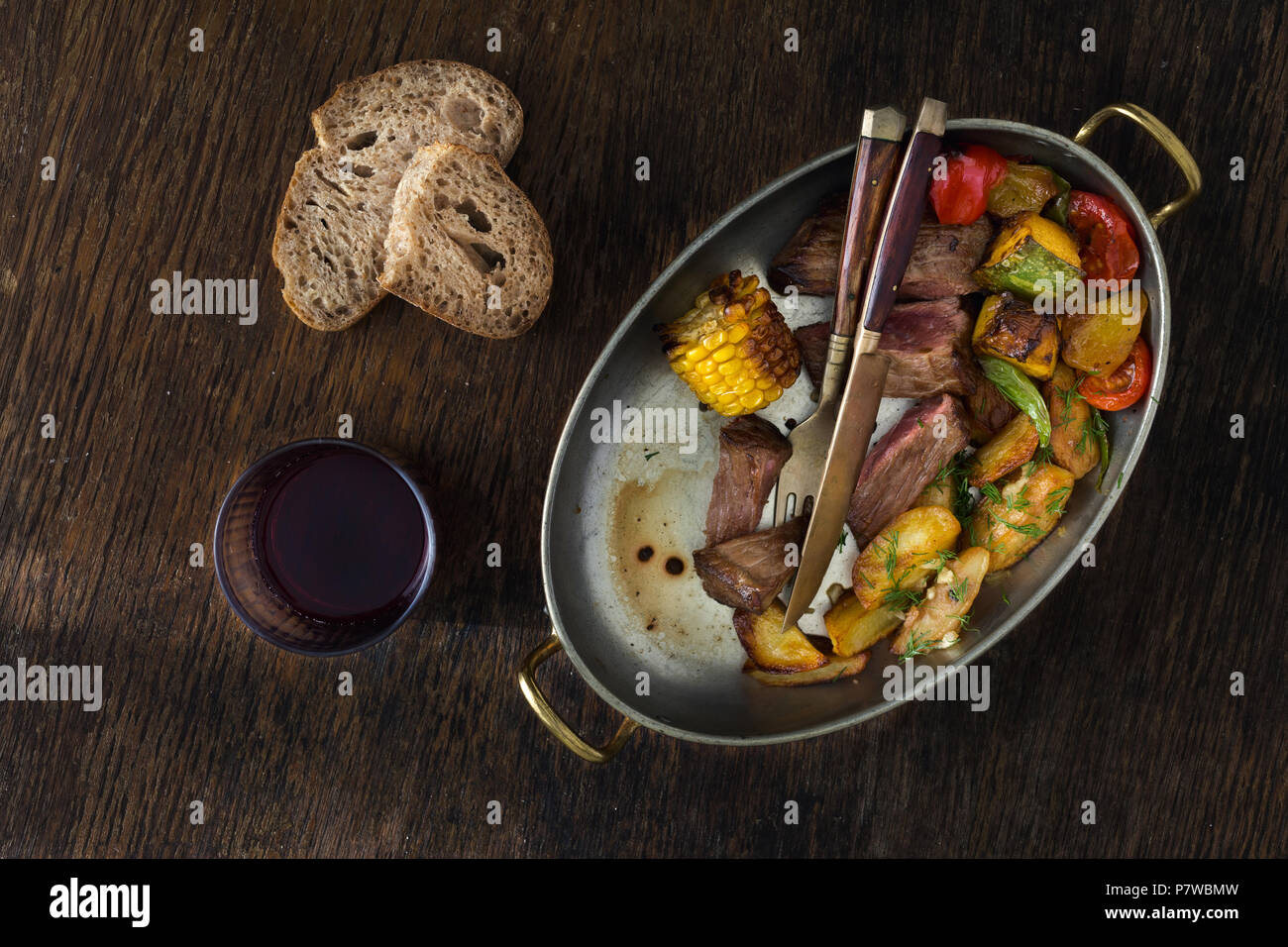 Carne filete con patatas fritas y verduras en sartén sobre fondo oscuro con un vaso de vino tinto, vista superior. Concepto de tabla de cena Foto de stock