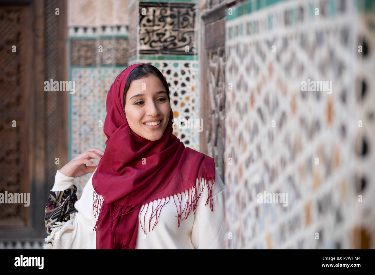 Retrato de mujer musulmana sonriente en trajes tradicionales con pañuelo rojo delante del muro decorado arabesco tradicional Foto de stock