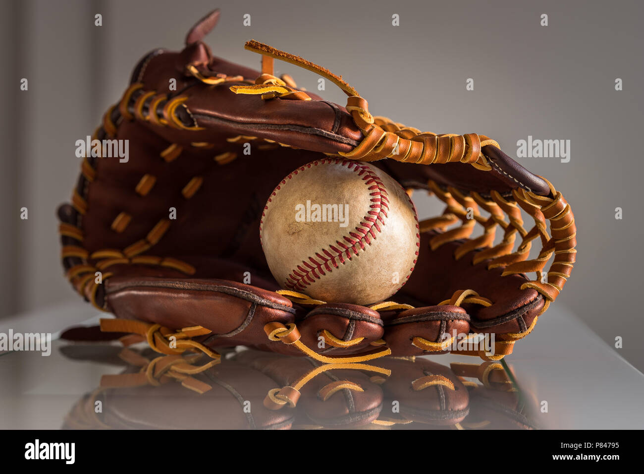 Close-up de una pelota de béisbol usado dentro de un guante de béisbol, cuero marrón sobre la llanura, fondo gris. Foto de stock