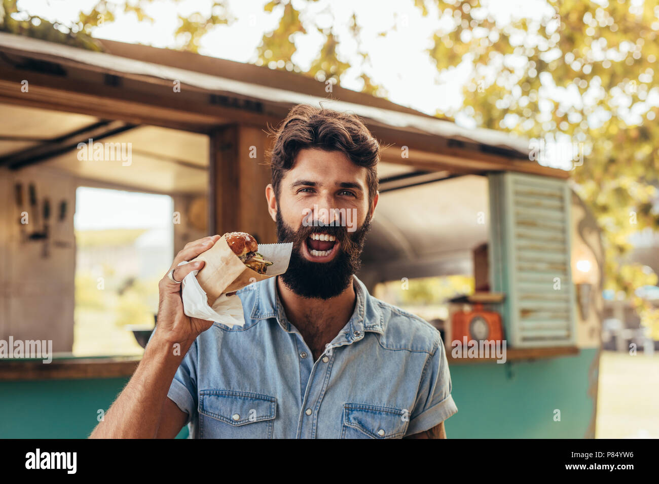 Muchacho joven con barba de comer una hamburguesa. El hombre tiene un delicioso burger afuera. Foto de stock