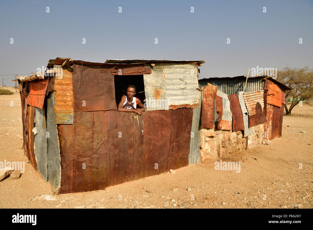 Hombre local mira por la ventana de su pobre choza de hierro corrugado, Spitzkoppe, región Erongo, Damaraland, Namibia Foto de stock