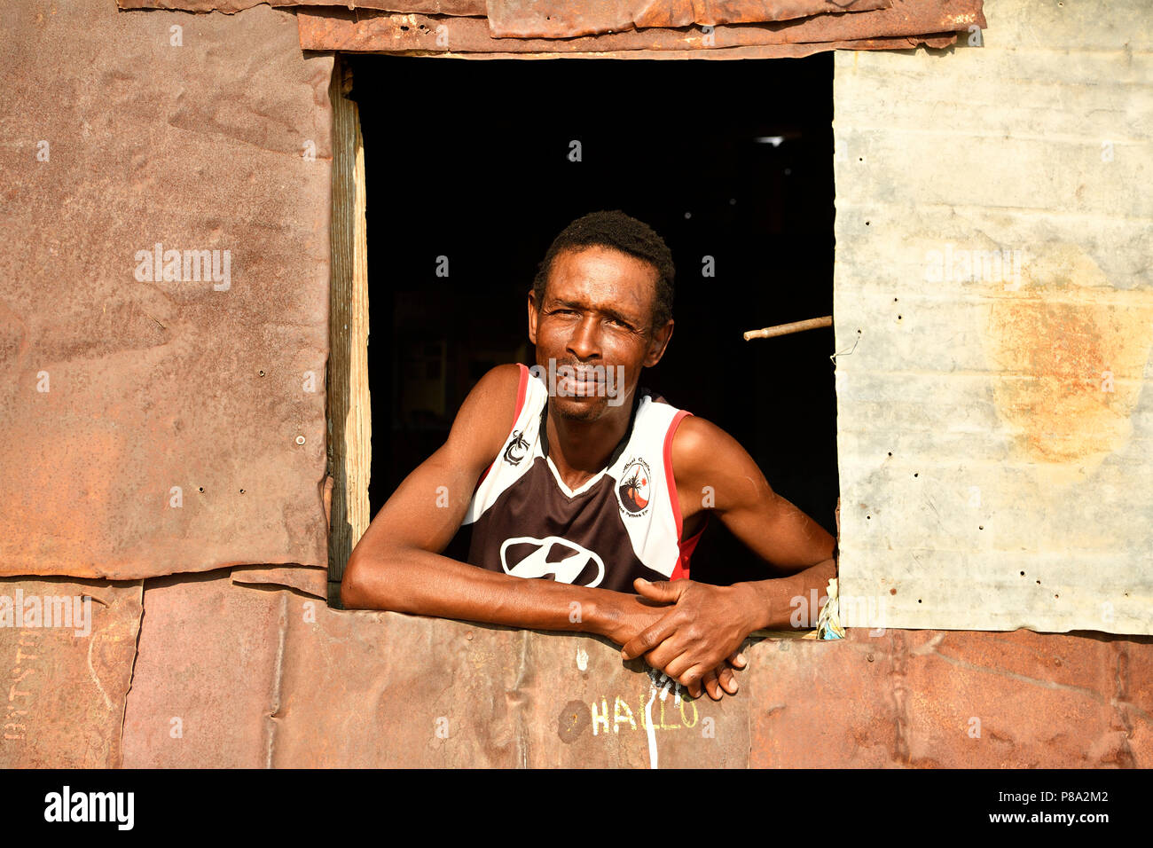 Hombre mirando por la ventana de su pobre choza de hierro corrugado, Spitzkoppe, región Erongo, Damaraland, Namibia Foto de stock