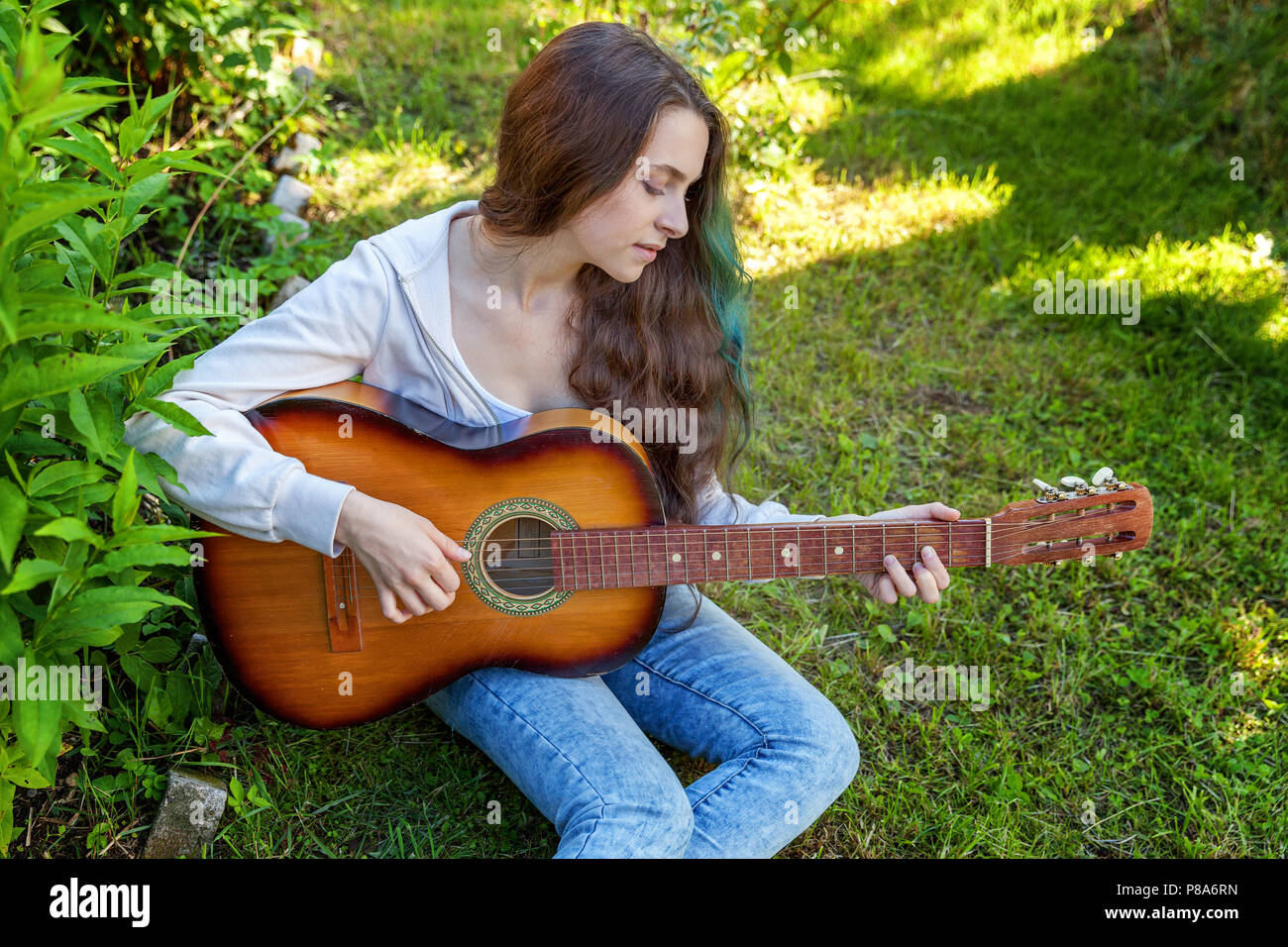 Young hipster mujer sentada en el césped y tocando la guitarra en el parque  o jardín de fondo. Jovencita aprendiendo a tocar la canción y escribir  música. Hobby, estilo de vida, instrumento,