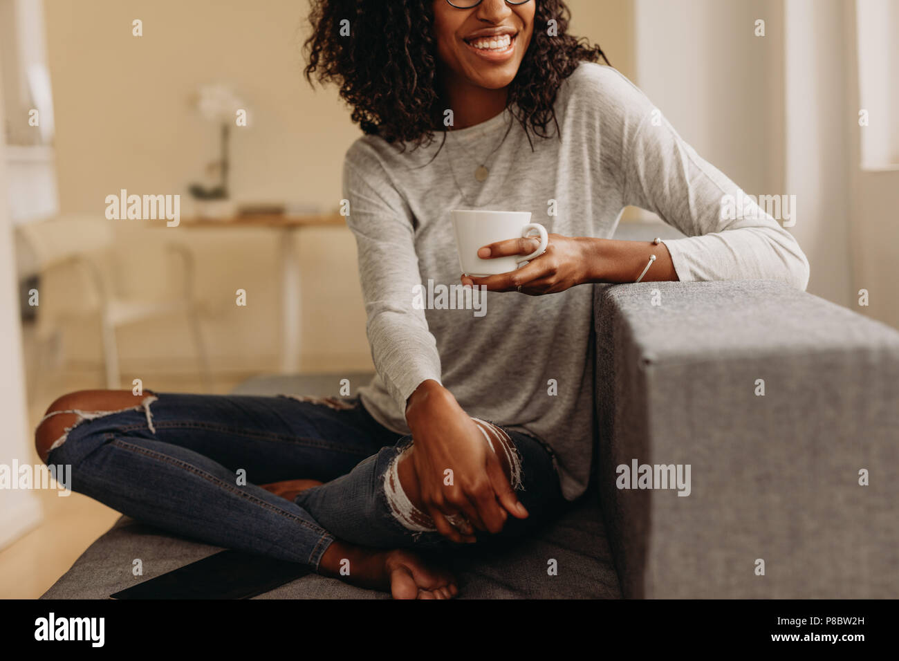 Mujer sonriente en moda jeans rasgados sosteniendo una taza de café para  relajarse en casa. Mujer sentada con las piernas cruzadas en el sofá en  casa bebiendo café y loo Fotografía de