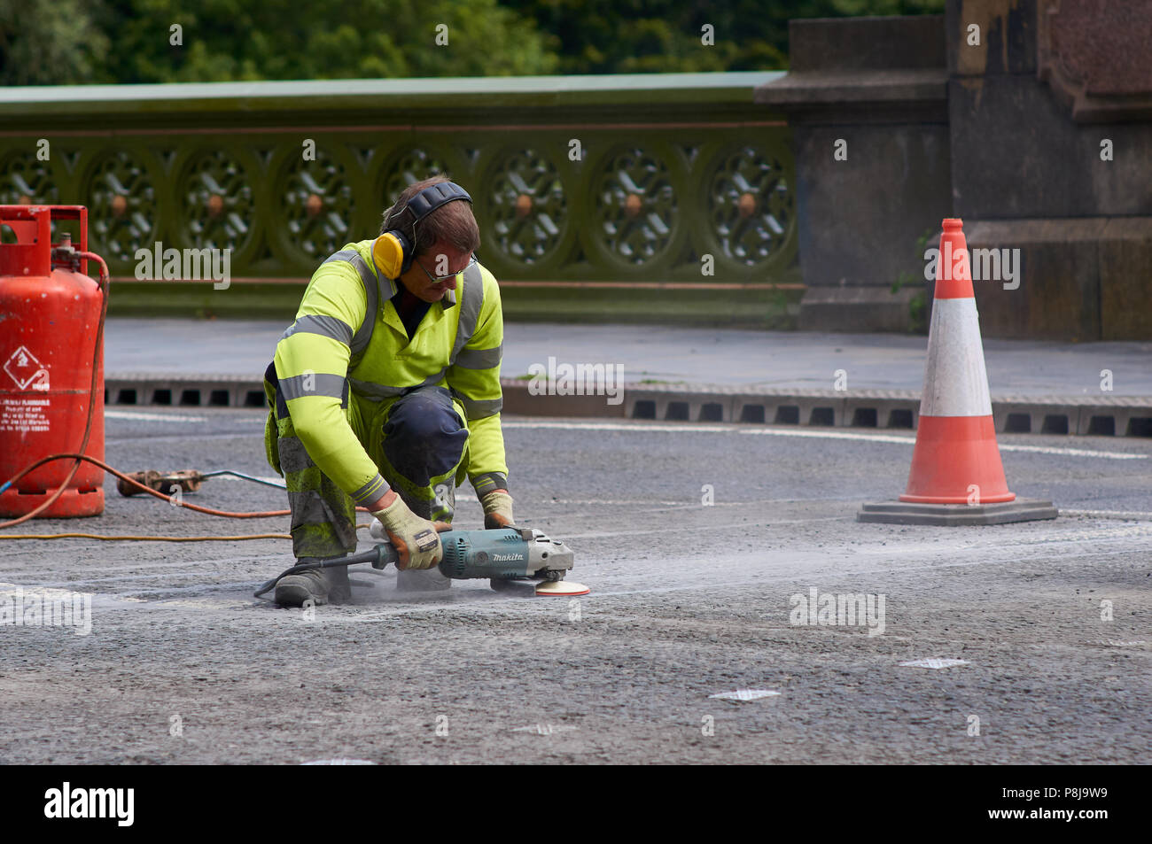 Un trabajador de mantenimiento vial mediante una amoladora de ángulo Power Tool para terminar las reparaciones de la superficie de la carretera. El trabajador es llevar orejeras y alta frente a la parte superior. Foto de stock