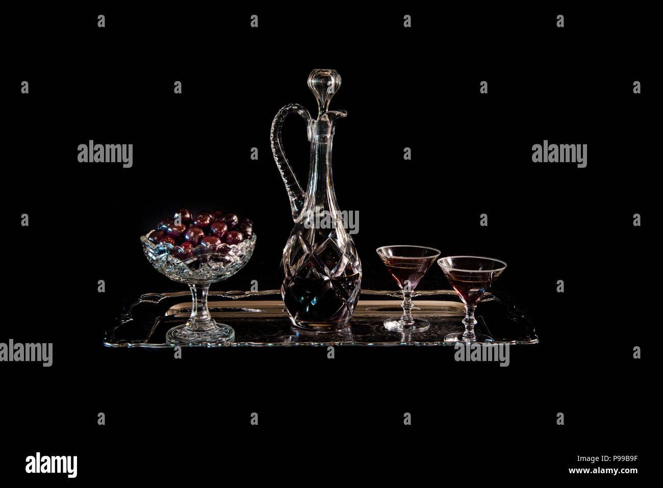 Las cerezas en un cuenco de cristal antiguo y una garrafa de Cristal y vasos con brandy cherry en un plato de plata con un fondo negro, un bodegón. Foto de stock