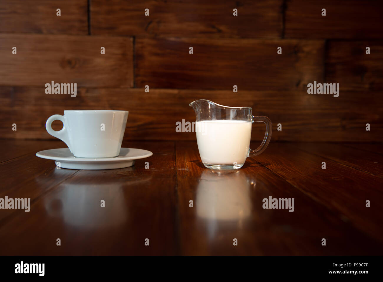 Leche en la jarra de leche de vidrio con taza blanca y el reflejo de una mesa de madera Foto de stock