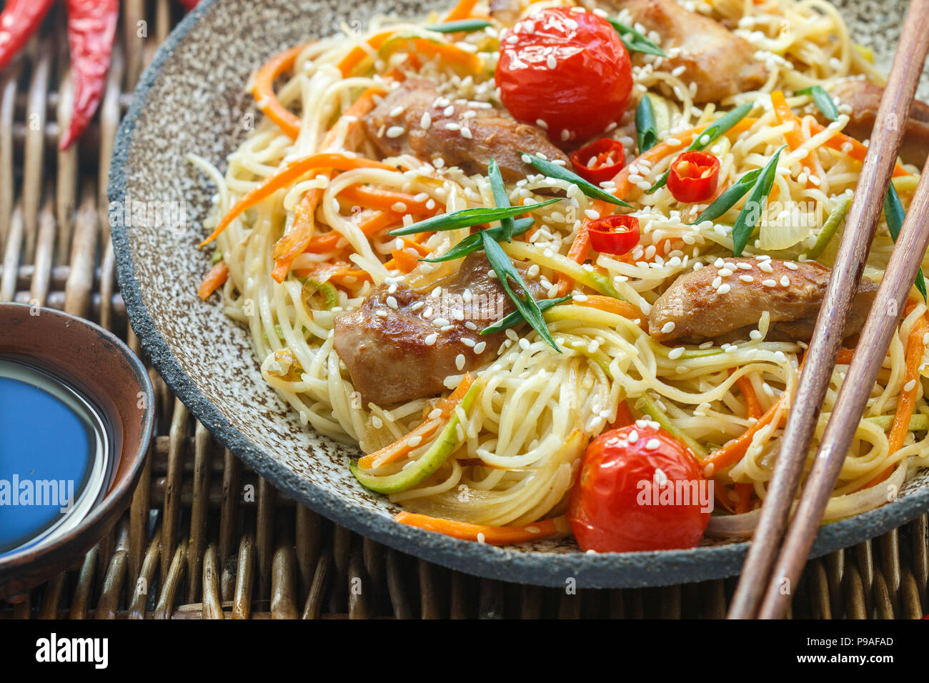 Revuelva de fideos fritos con pollo, hortalizas (tomate, zanahoria, calabacín, ajo, pimiento, cebolla verde), semillas de sésamo y salsa de soja en el cierre de la placa de arcilla Foto de stock