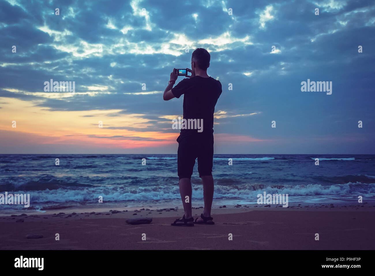 Silueta de hombre conservar recuerdos con la cámara del teléfono en su mano. Maravillosa puesta de sol en el mar, buen nivel de agua Foto de stock