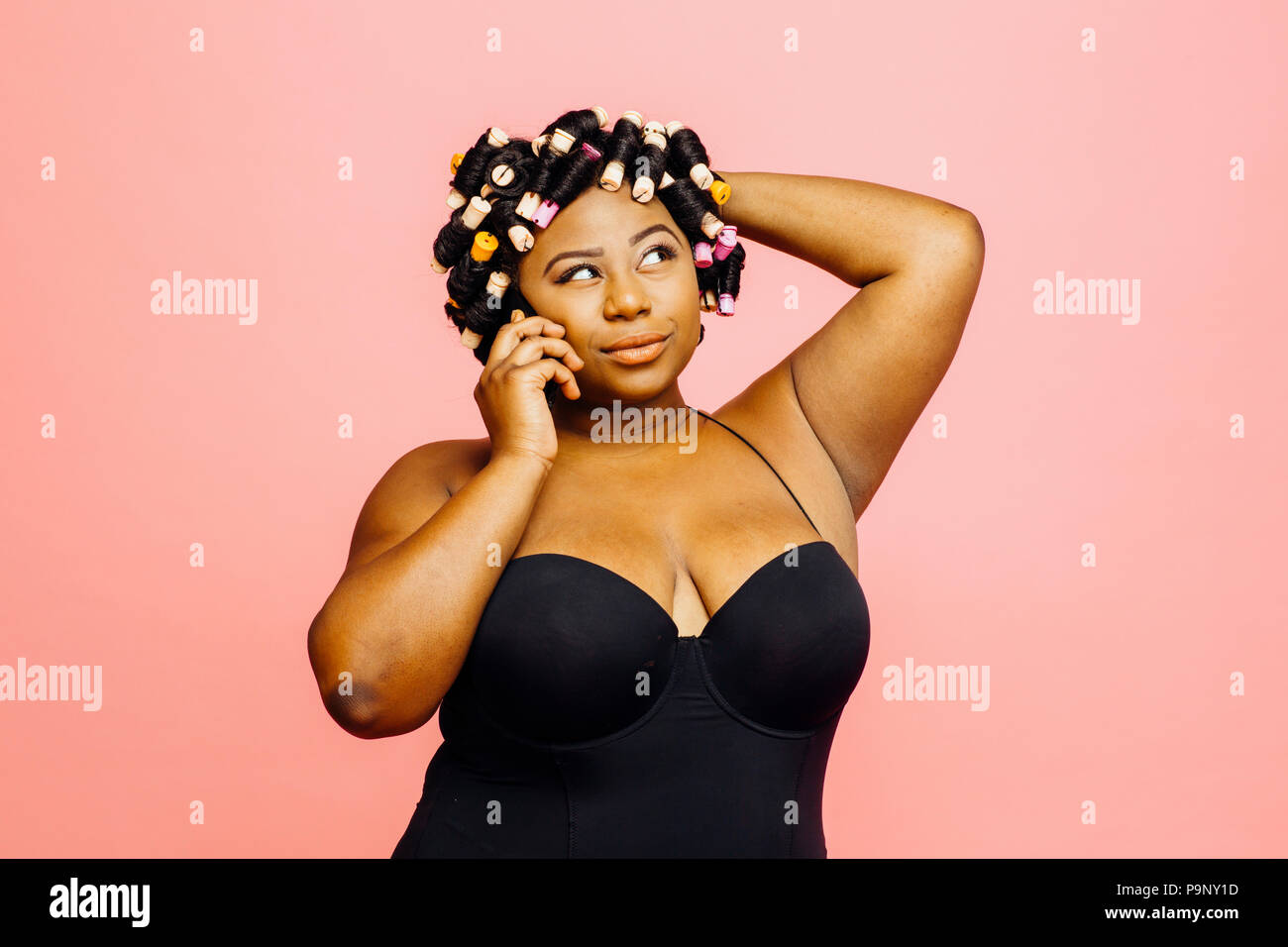 Mujer con rizadores de pelo y neglige hablando por el teléfono y mirando hacia arriba, aislado sobre fondo de color rosa Foto de stock