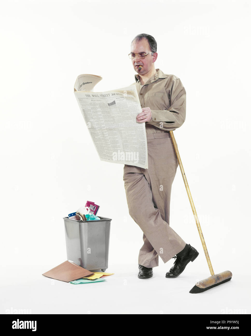 1970 CONSERJE HOMBRE DE MANTENIMIENTO apoyándose en escoba Cigarro fumar  leyendo el periódico WALL STREET JOURNAL POR OFFICE PAPELERA - ks11079  HAR001 HARS COPIA ESPACIO PERSONAS DE LONGITUD COMPLETA ESCOBA hombres  residuos
