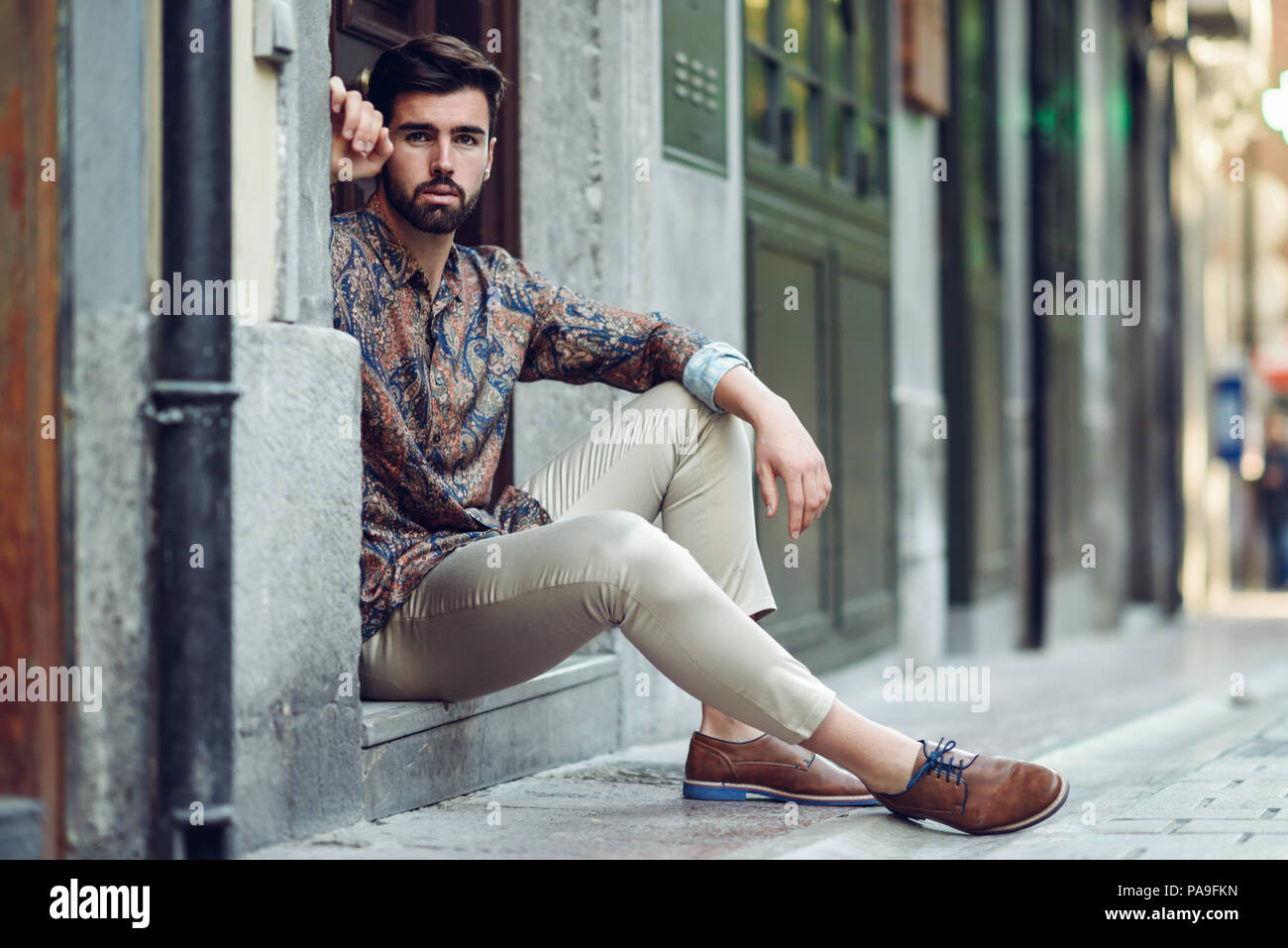 Joven Hombre Barbado, modelo de moda sentados en un paso vistiendo ropa casual. Chico con barba peinado moderno mirando a otro lado de la calle Fotografía de stock - Alamy