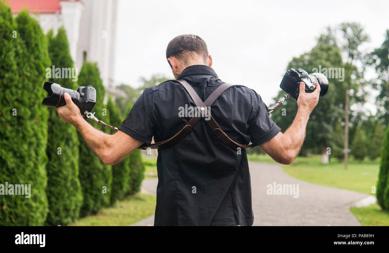Fotógrafo profesional de bodas en camisa negra y con tirantes para los  hombros sosteniendo cámaras como un fusiles contra el jardín verde.  Fotografía de la boda Fotografía de stock - Alamy
