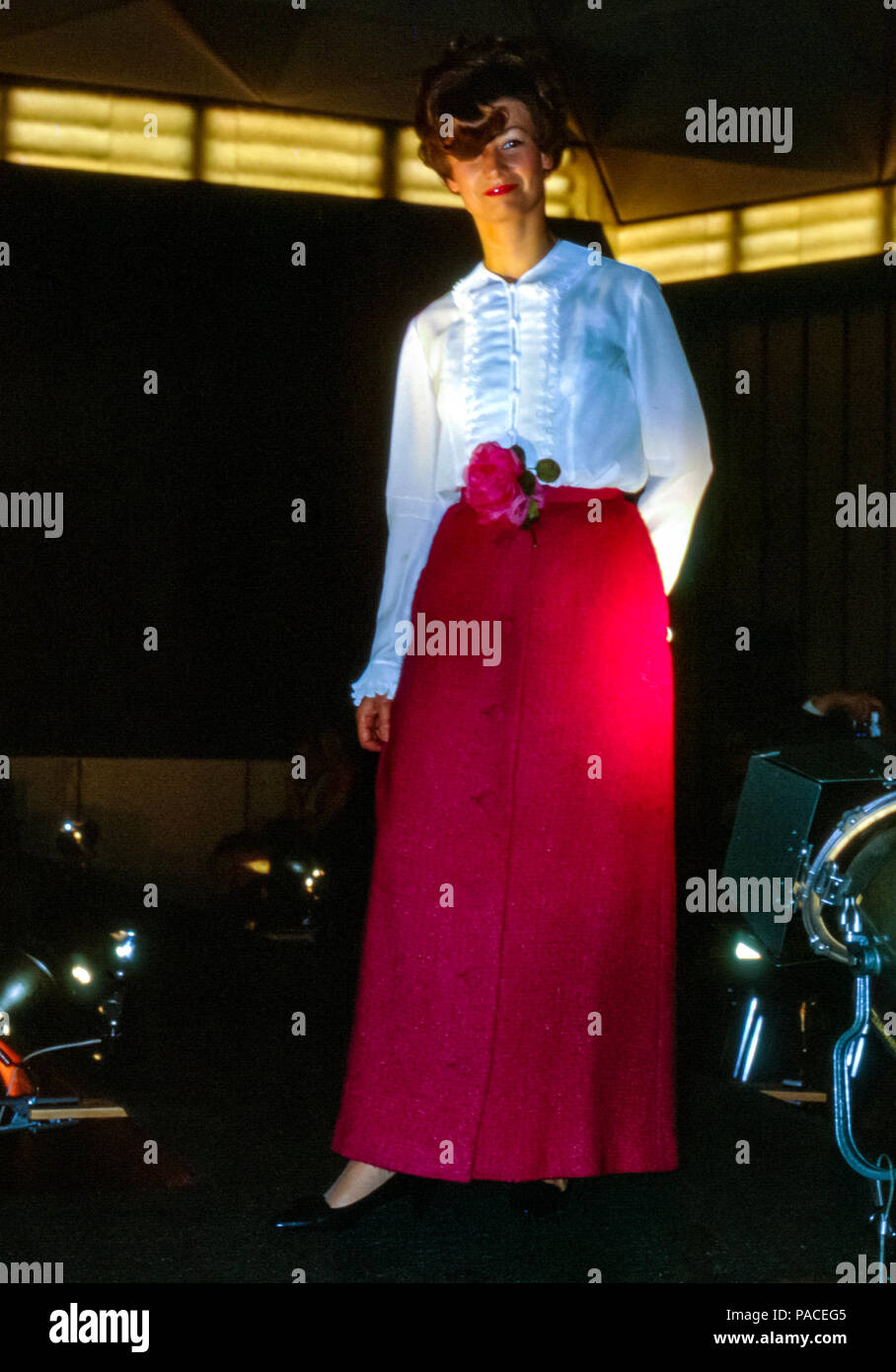 Mujer atractiva en una pasarela modelando ropa de noche formal con una falda  roja larga y blusa blanca frilly, en un desfile de moda amateur, Hamburgo,  Alemania en la década de 1960