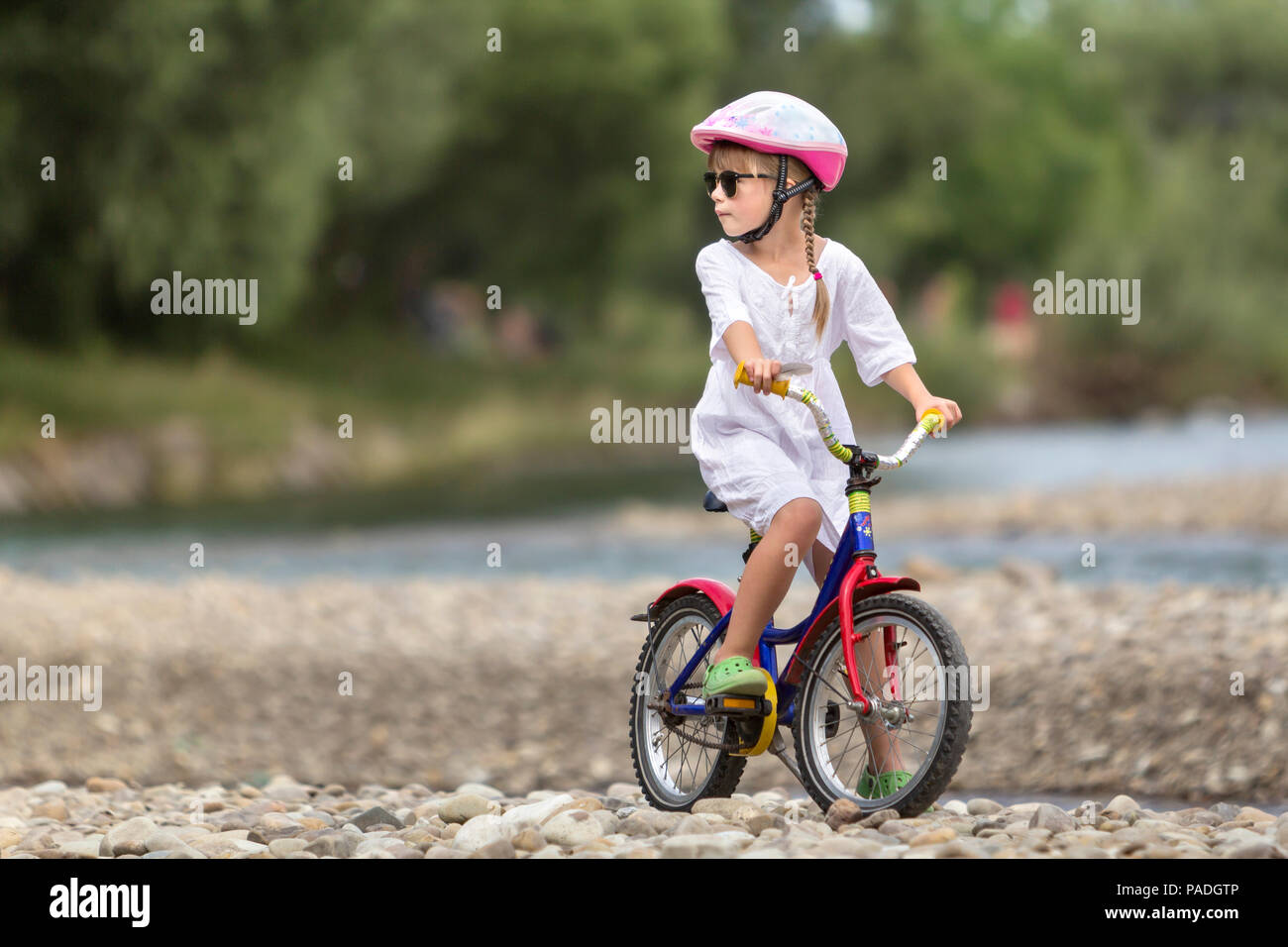 Linda jovencita en ropa blanca, gafas de sol con largas trenzas llevar casco de seguridad rosa montando bicicleta infantil de guijarros de río verde borrosa Foto de stock