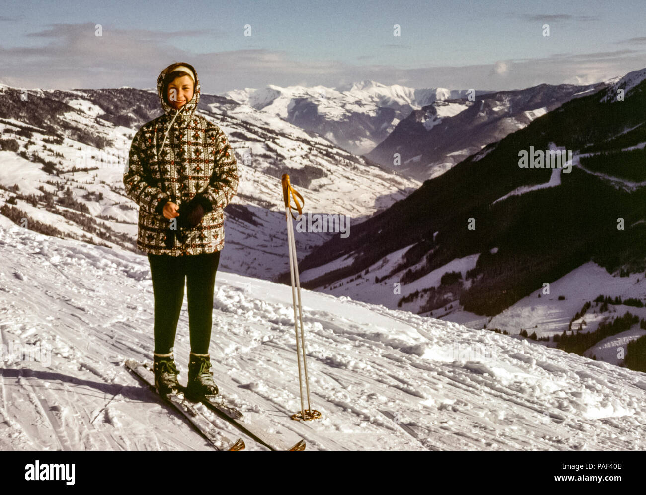 Mujer con botas de esquí de cuero y esquís posando en la pista de esquí de  la cima de la montaña en Hinterglemm, Austria con vistas a un valle en la  década