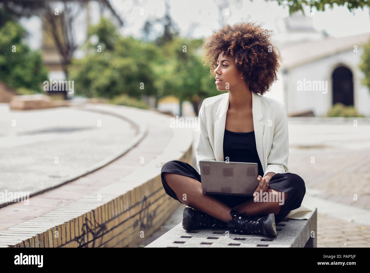 Moda joven con el cabello rizado sentado en un banco con portátil Foto de stock