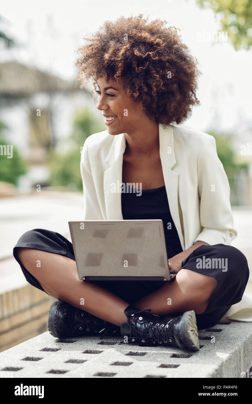 Moda joven con el cabello rizado sentado en un banco con la laptop riendo Foto de stock