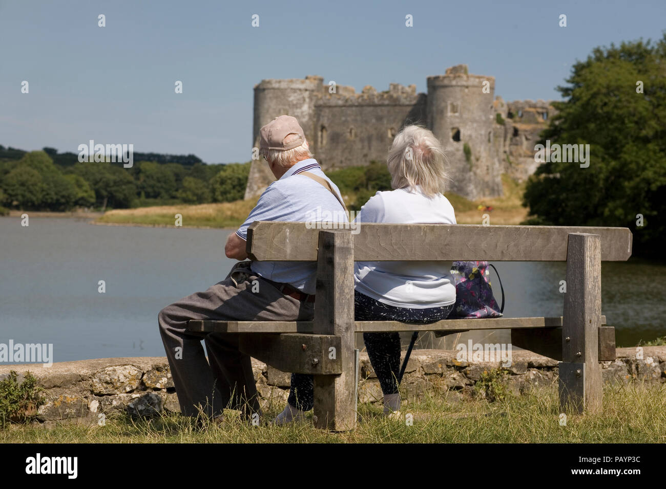 Pareja de ancianos sentados en un banco por Carew tidal piscina mirando hacia Carew Castle en una calurosa tarde de verano Foto de stock