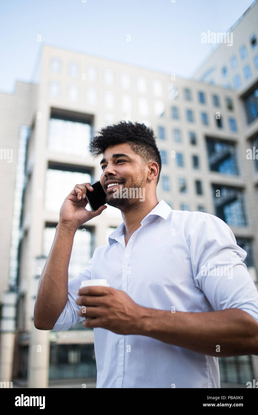Joven apuesto hombre afro americana de pie delante del enorme edificio moderno de negocios, sonriente, sosteniendo el café para ir y hablar por teléfono móvil. Foto de stock