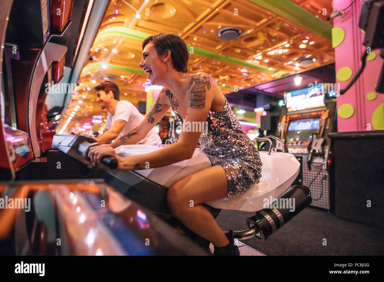 Feliz pareja divertirse jugando juegos de carreras arcade en un salón de juegos. Emocionados mujer jugando un juego de carreras arcade sentado en una bicicleta de carreras. Foto de stock
