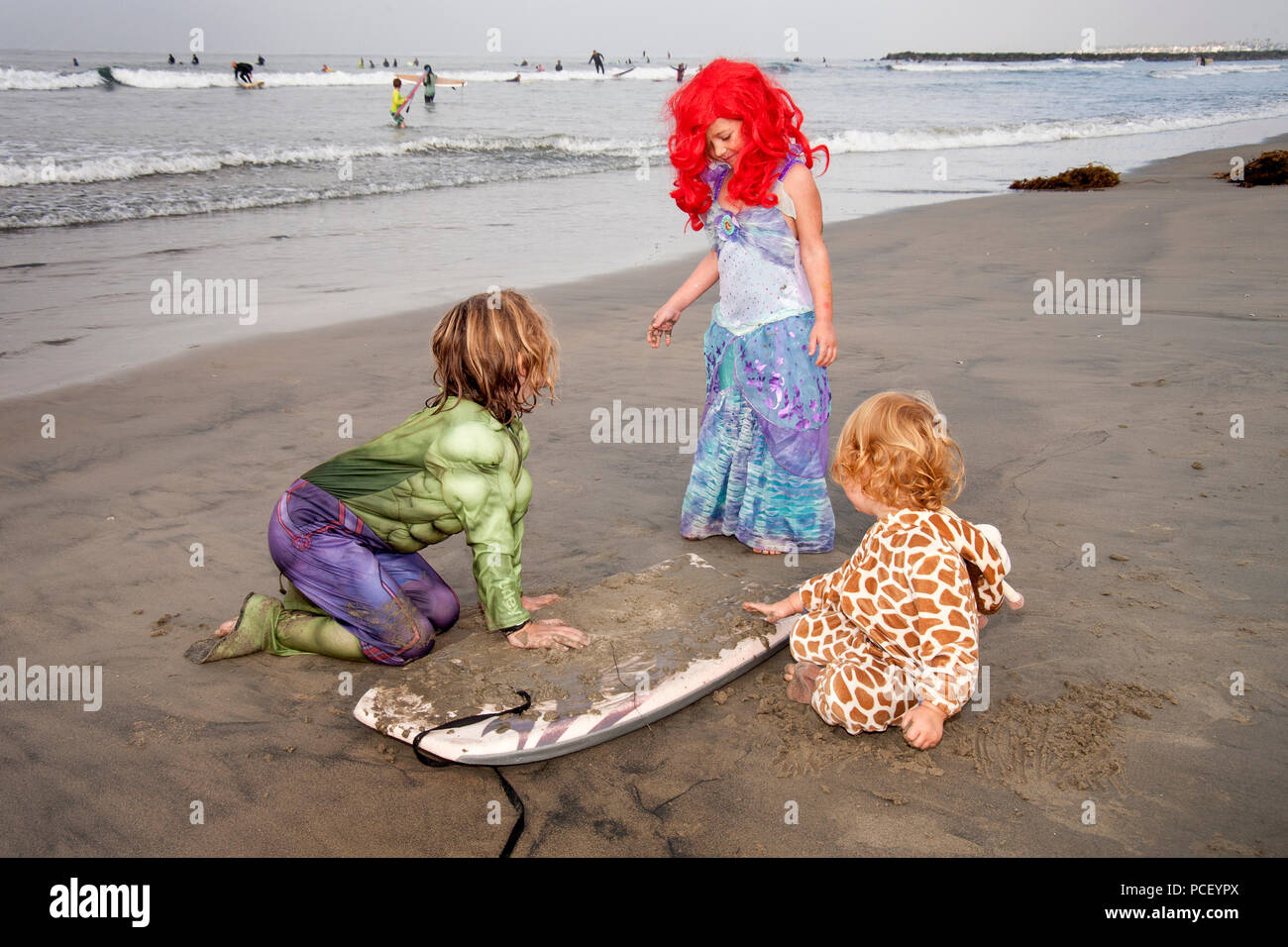 Vestida con un traje de Hulk, un niñito sands su tabla de surf junto con niñas  vestidas como una sirena y una jirafa en un evento de disfraces de  Halloween de surf