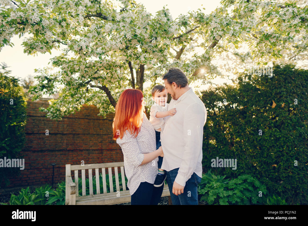 Hermoso y elegante redhead mom en una blusa blanca de pie con su bonito  hombre vestido con una camisa blanca y pantalones de mezclilla; juegan con  sus litt Fotografía de stock -