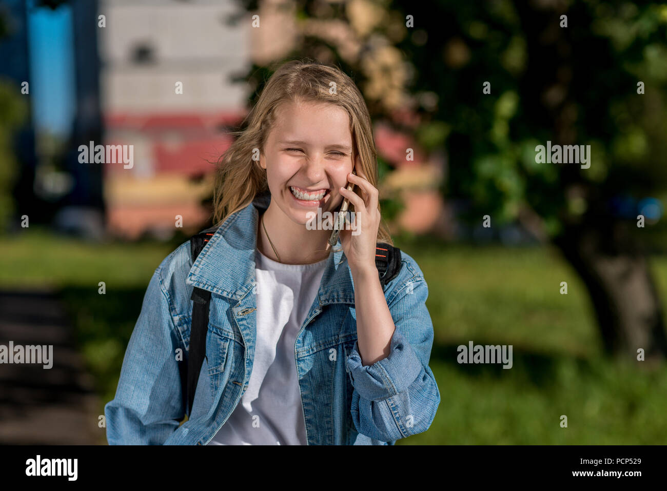 Chica colegiala hablando por teléfono, en verano en la naturaleza, sonriendo felizmente, emocionalmente feliz. En sus manos sostiene un smartphone, en jeans chaqueta. Foto de stock