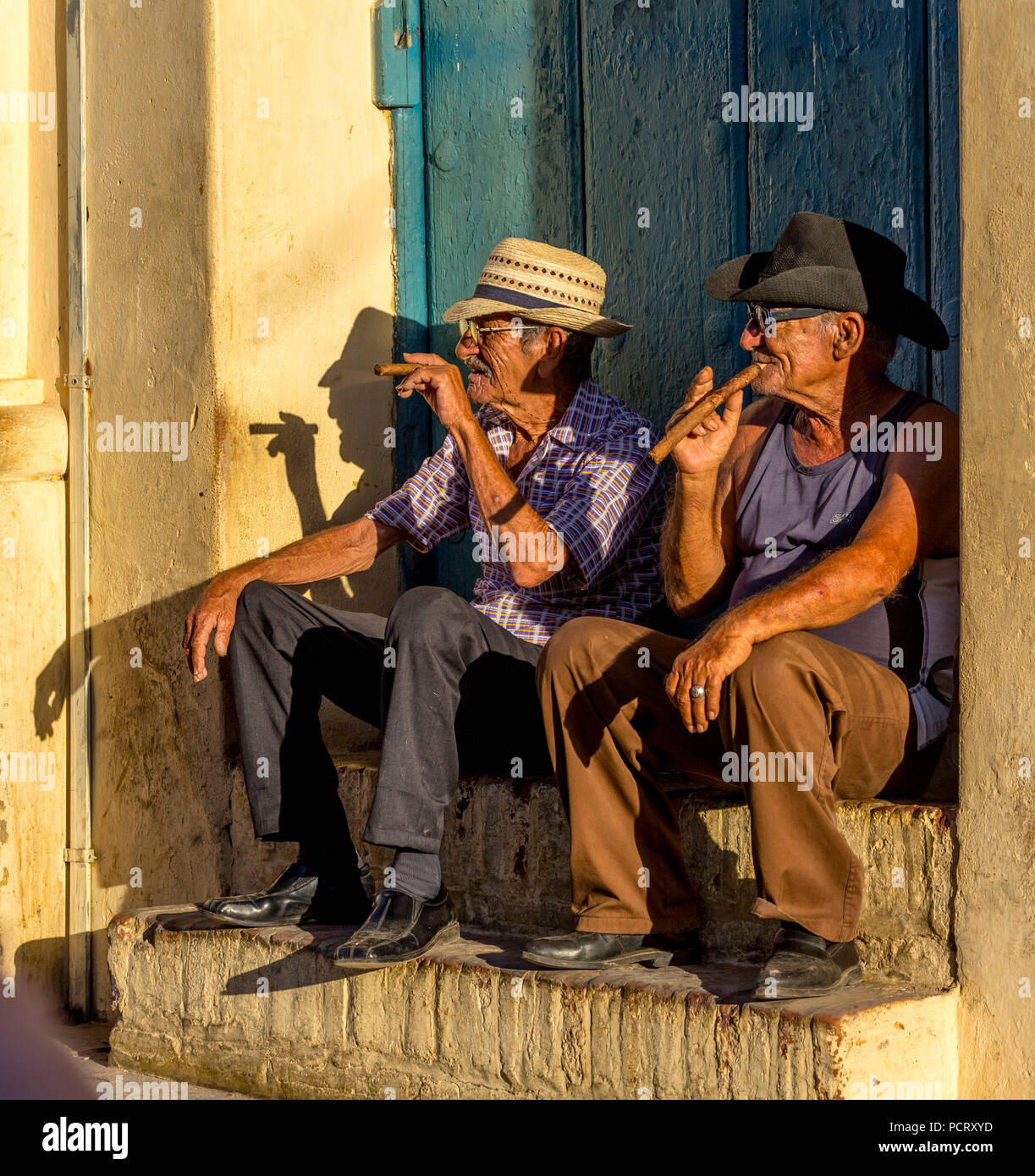 Dos viejos cubanos fuman cigarros y sentarse en una escalera en la cálida luz del atardecer, Trinidad, Cuba, Sancti Spíritus, Cuba Foto de stock