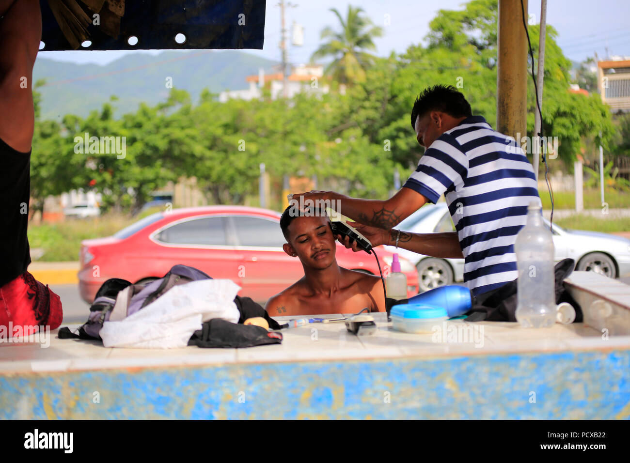 Puerto La Cruz, Anzoátegui, Venezuela. 3 ago, 2018. Agosto 03, 2018. Un  hombre realiza cortes de pelo, en una improvisada calle Barber shop, en el  mercado de cocos, en la ciudad de