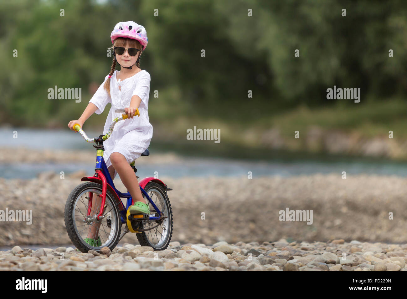 Linda jovencita en ropa blanca, gafas de sol con largas trenzas llevar casco de seguridad rosa montando bicicleta infantil de guijarros de río verde borrosa Foto de stock