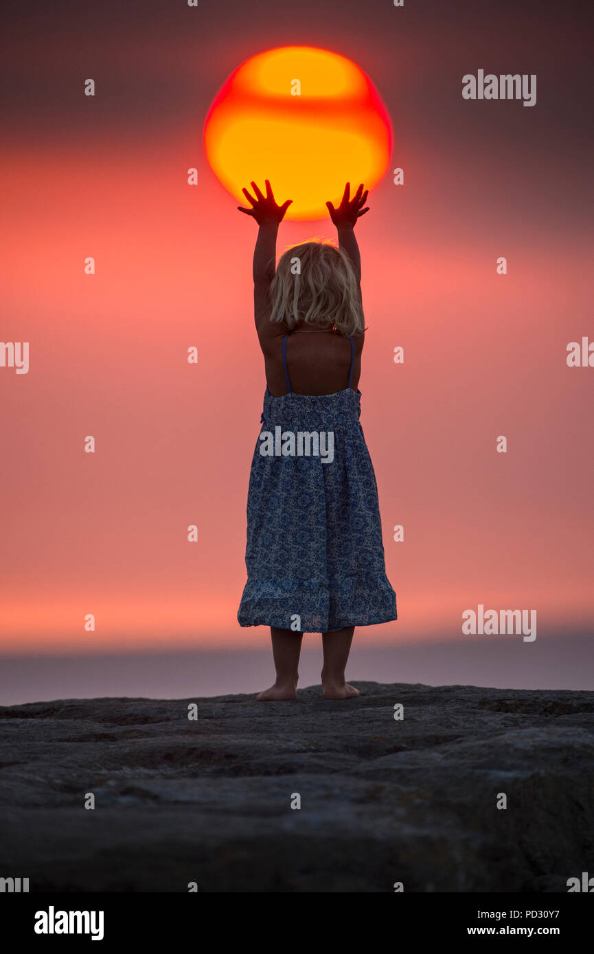 Niña alcanzando hasta tocar la puesta de sol, vista trasera, perspectiva falsa, Doolin, Clare, Irlanda Foto de stock