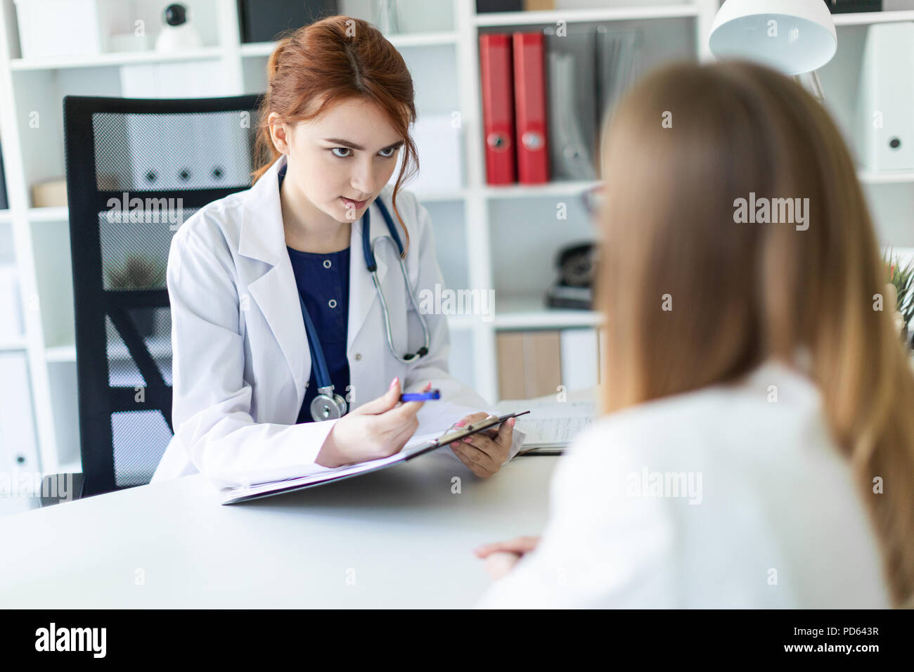 Una hermosa joven vestida con una túnica blanca está sentado en el escritorio de la oficina y la comunicación con el interlocutor. La niña toma notas en la hoja. Foto de stock