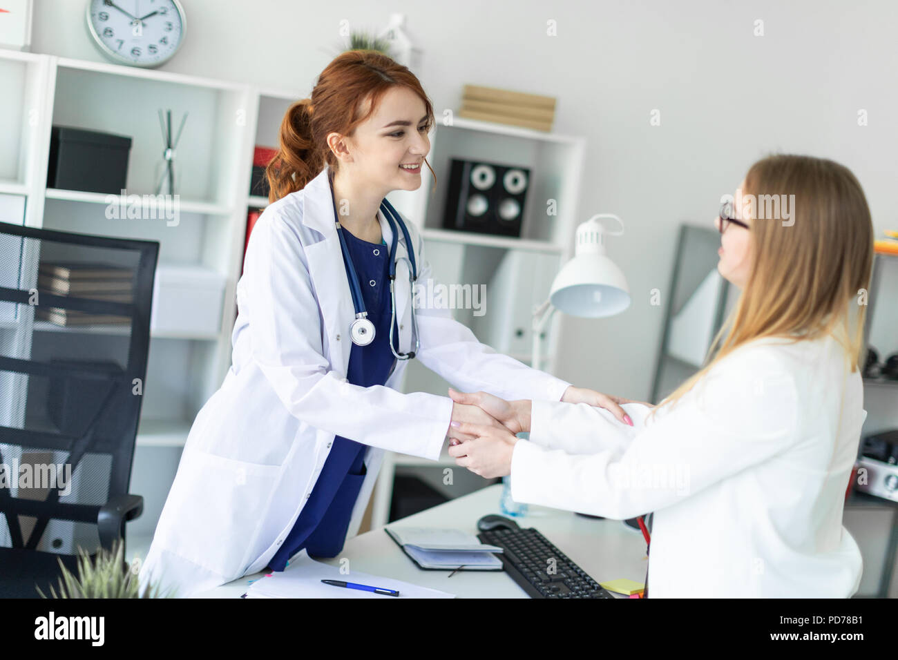 Una hermosa joven en una bata blanca está de pie cerca de la mesa en la oficina y un apretón de manos con el paciente. El paciente gracias al doctor. Foto de stock